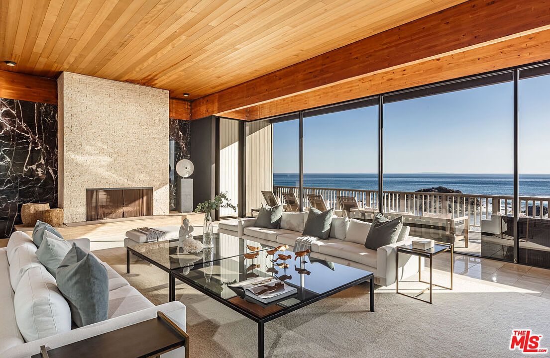 This is a luxurious living room featuring a modern fireplace with stone and marble accents, a sleek glass coffee table, and a large white sofa with stylish pillows. Floor-to-ceiling windows offer stunning ocean views, creating a bright and airy atmosphere. The wood-paneled ceiling adds warmth and character to the space.