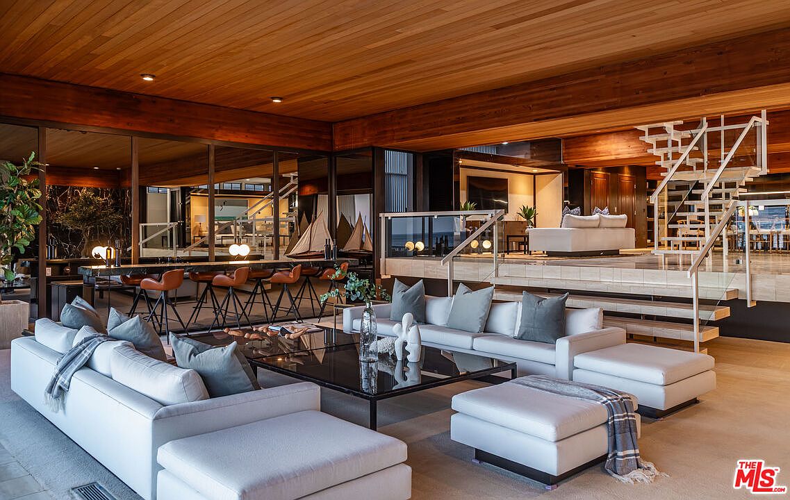 This is a spacious living room featuring a modern design with a warm, inviting atmosphere. The room includes multiple white sofas with gray pillows, a dark glass coffee table, and a unique white staircase. The wooden ceiling and beams add a touch of rustic elegance, while the large windows provide ample natural light and views of the surrounding area.