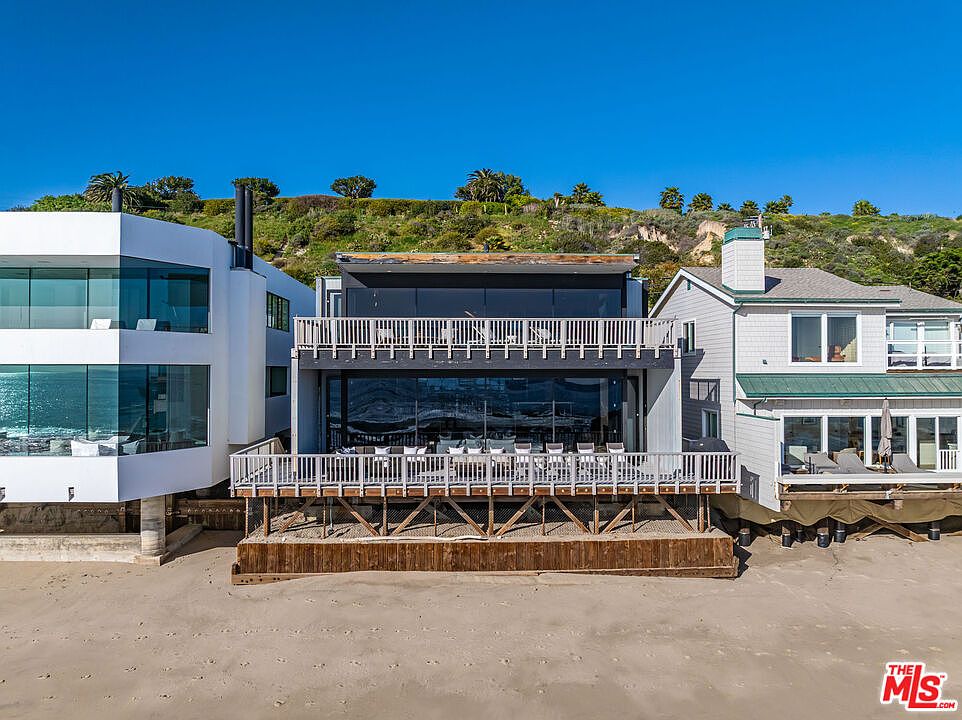 This is a rear view of three beachfront houses. The center house has a dark gray exterior with large glass windows and white railings on the balconies. The house on the left is white with large windows, and the house on the right is gray with a green roof. The houses are built on stilts above the sand.