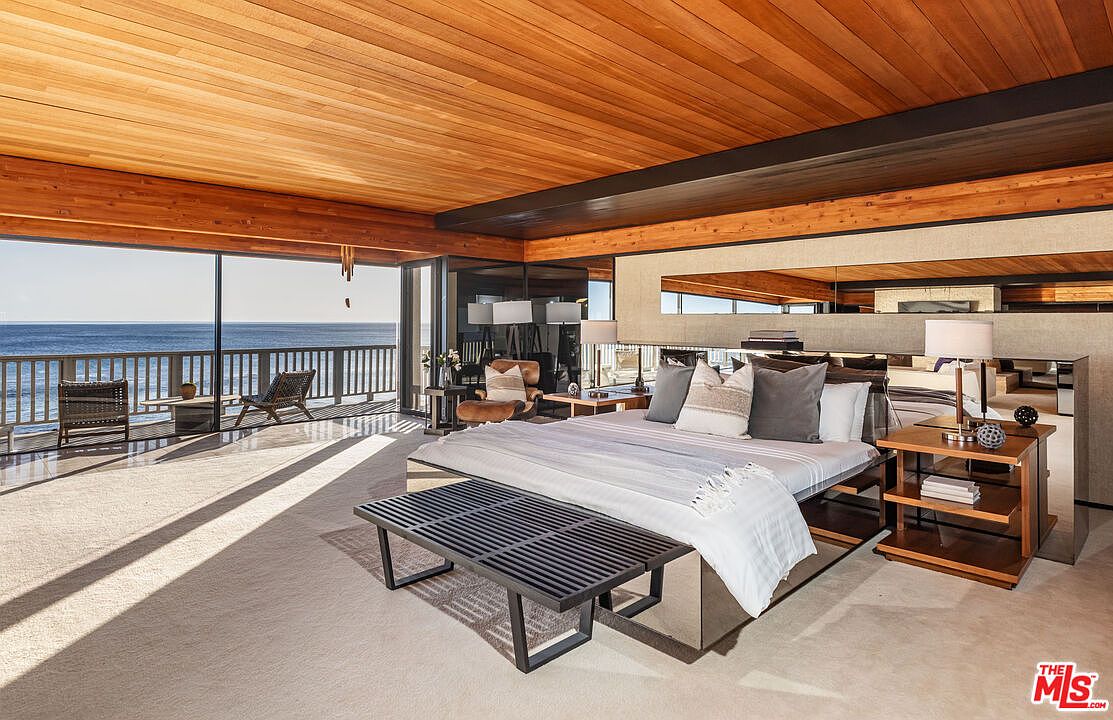 This is a luxurious primary bedroom with a stunning ocean view. The room features a wood-paneled ceiling, a mirrored bed frame, and a private balcony. The neutral color palette and minimalist decor create a serene and sophisticated atmosphere, perfect for relaxation.