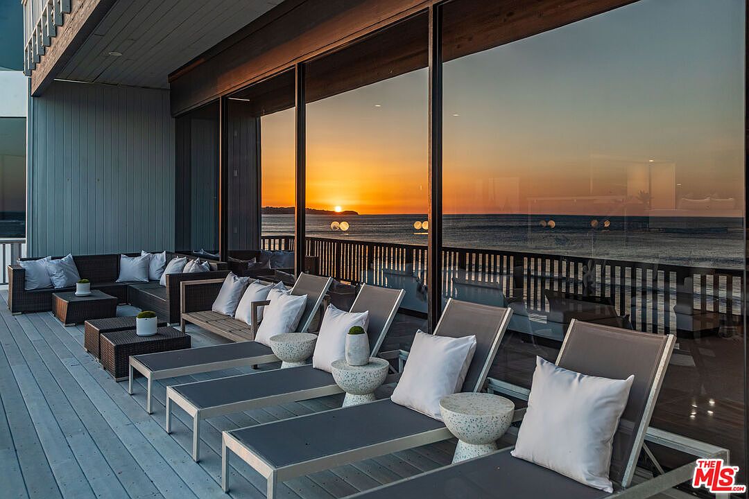 This image showcases a luxurious outdoor patio or balcony area, featuring modern lounge furniture including chaise lounges and a sectional sofa with throw pillows. The space is designed for relaxation and entertainment, with small side tables and decorative plants adding to the ambiance. Large glass windows offer stunning ocean views at sunset, enhancing the property's appeal.