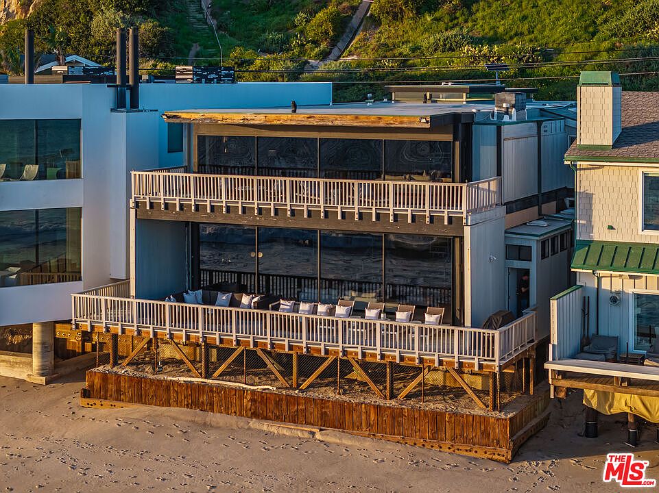 This image showcases the rear exterior of a multi-level beachfront property. The building features large windows offering ocean views, multiple balconies with white railings, and a wooden support structure at the base. The overall impression is one of luxury and coastal living.