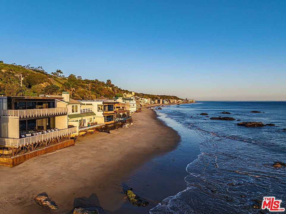 This aerial view showcases a row of beachfront homes nestled along a sandy shore, with the ocean stretching out to the horizon under a clear blue sky. The homes feature balconies and decks, offering stunning ocean views, while the beach provides direct access to the water. The overall impression is one of luxury coastal living.