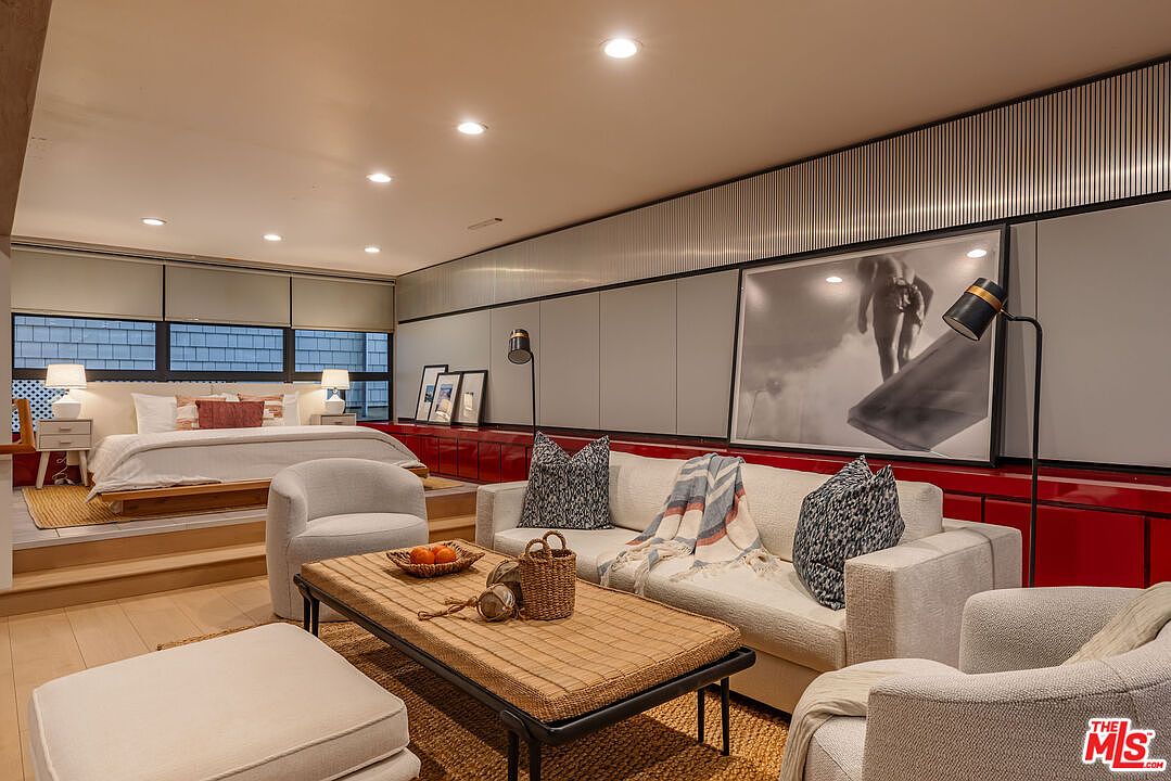This interior shot showcases a combined living and bedroom space, featuring a raised platform for the bed and a cozy living area with a sofa, armchairs, and a woven coffee table. The room is decorated with modern art and stylish accessories, creating a sophisticated and inviting atmosphere. The color palette is neutral with pops of red.