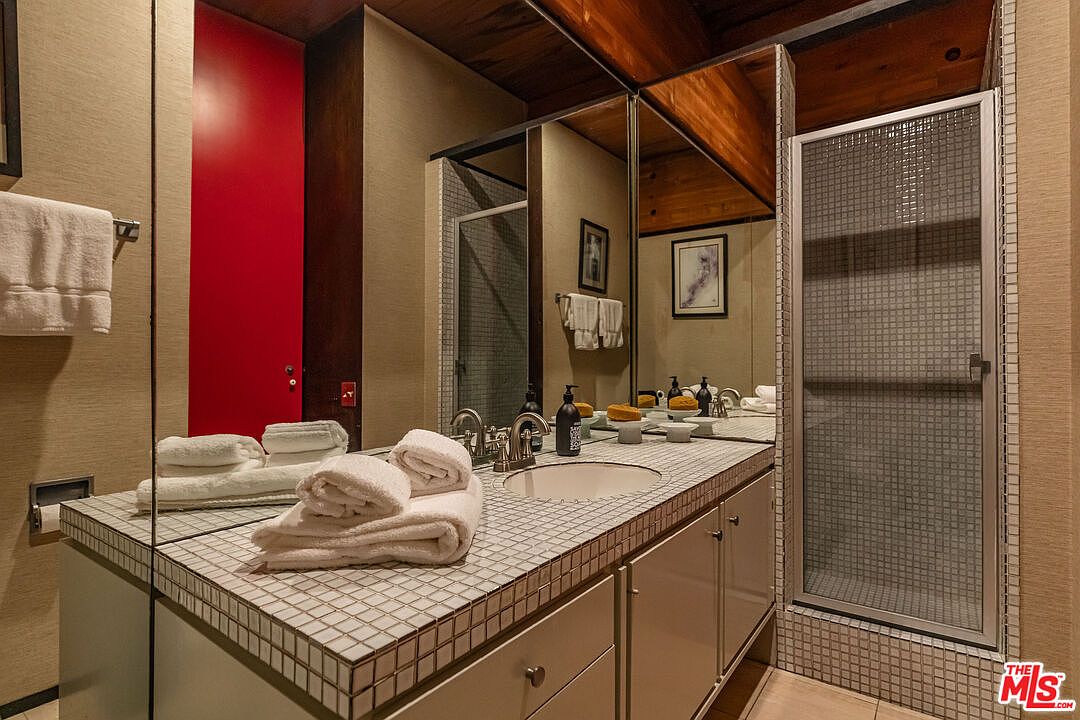This is a primary bathroom featuring a unique tiled countertop with a built-in sink and a mirrored backsplash. A red door adds a pop of color, while a glass-enclosed shower is visible to the right. The bathroom has a retro style with wood ceilings and neutral wall tones.