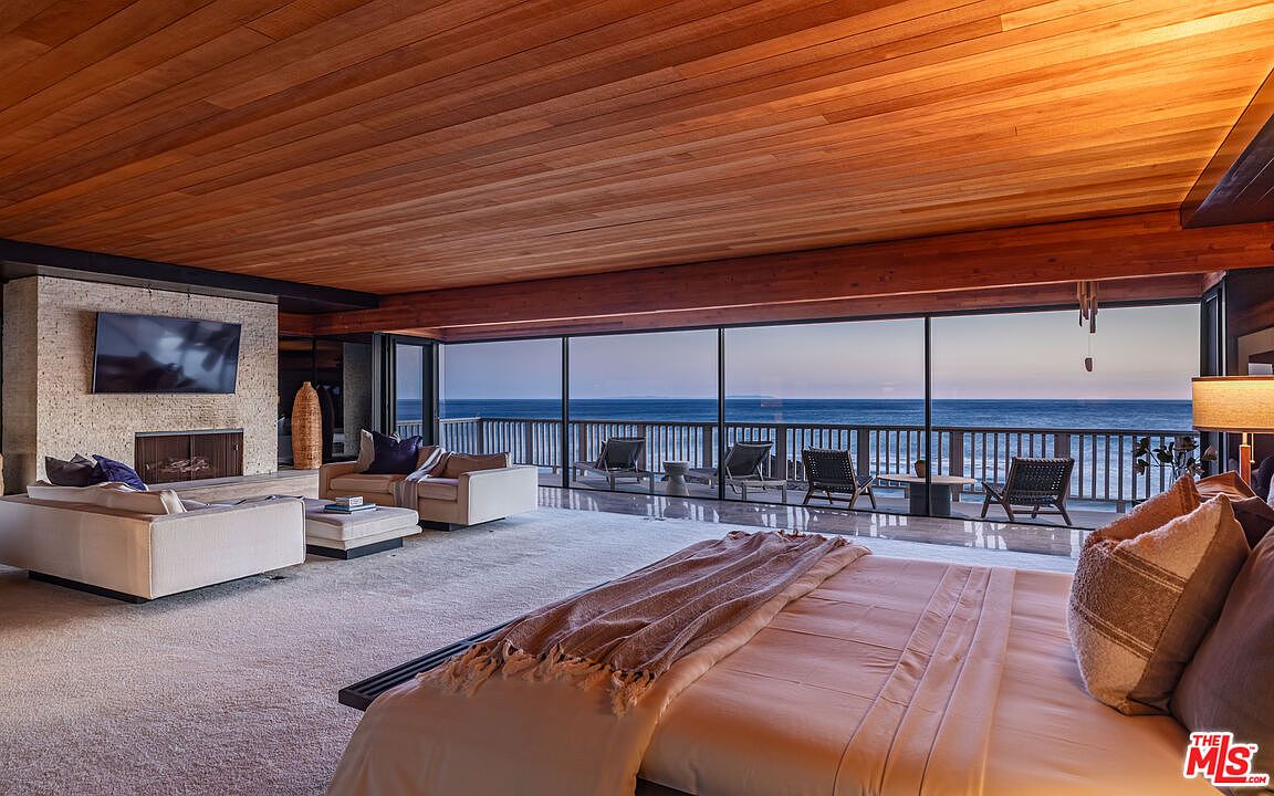 This is a luxurious primary bedroom with a stunning ocean view. The room features a wooden plank ceiling, a stone fireplace with a mounted TV, and a seating area with modern sofas. The bed is prominently displayed in the foreground, and the large windows offer an unobstructed view of the ocean and a balcony with lounge chairs.