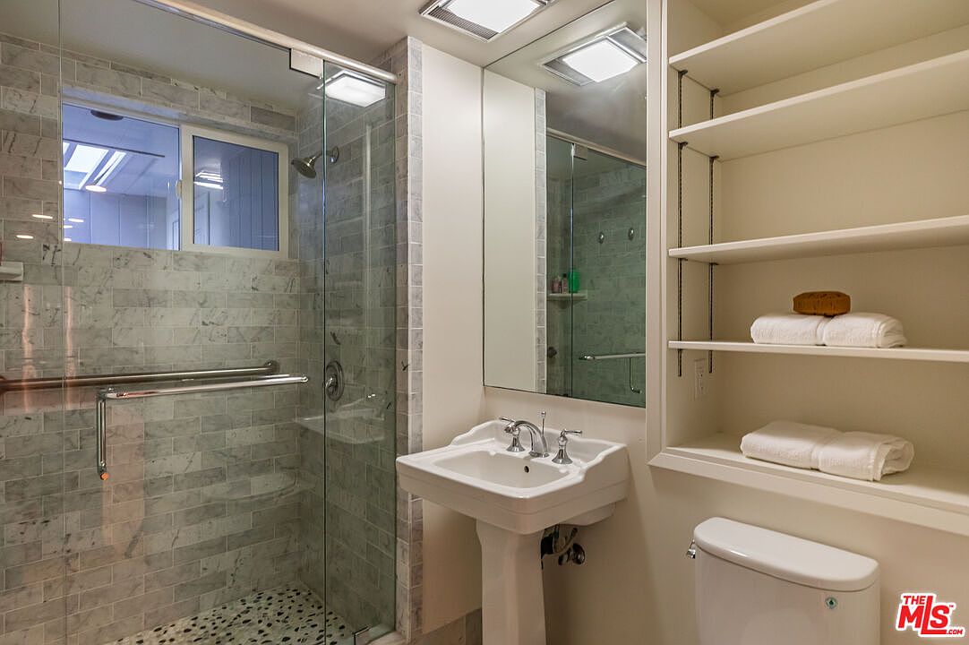This is a well-lit bathroom featuring a glass-enclosed shower with gray marble tiling and a pebble floor. A pedestal sink with chrome fixtures sits next to a built-in shelving unit, which is adjacent to the toilet. The overall impression is clean and modern.