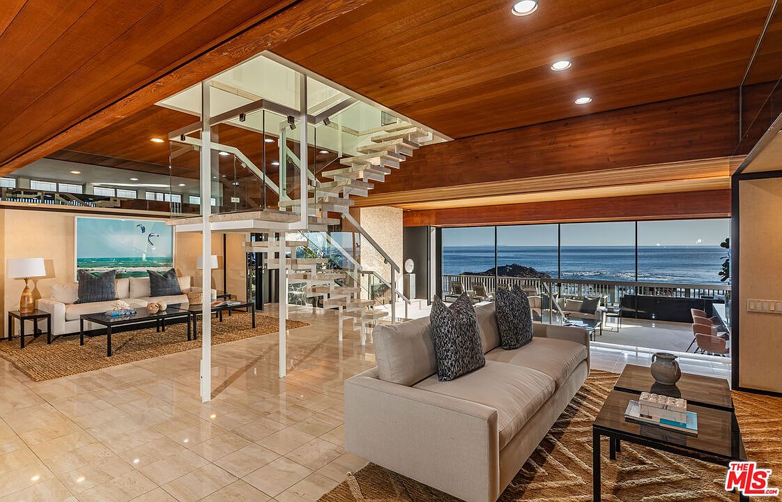 This is an interior shot of a luxurious living room featuring a unique architectural staircase with glass railings. The room is well-lit with natural light streaming in from large windows that offer ocean views. The space is furnished with comfortable sofas and stylish coffee tables, creating an inviting and sophisticated atmosphere.