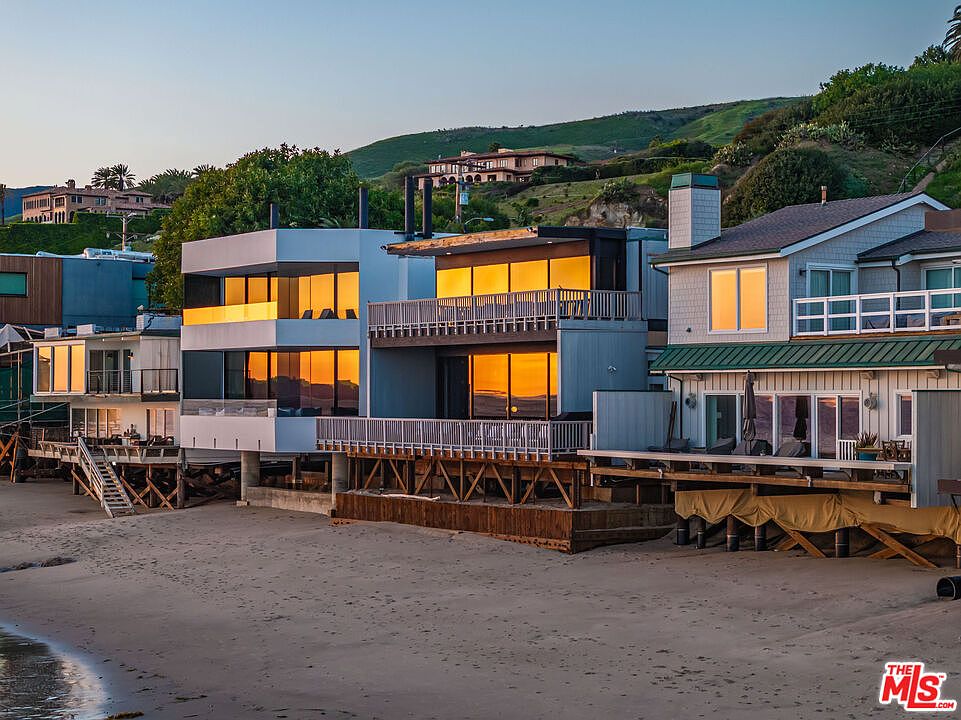 This image showcases the rear exterior of several beachfront homes, highlighting their decks and balconies overlooking the sandy beach. The architecture varies, with modern, boxy structures alongside more traditional, shingled designs. The setting sun casts a warm glow on the windows, creating an inviting and luxurious coastal living atmosphere.
