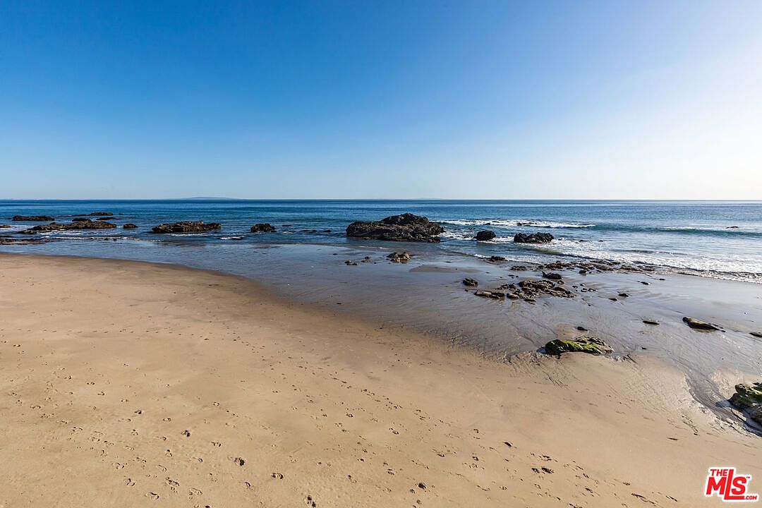 This image showcases a serene beach scene with a clear blue sky meeting the ocean horizon. The sandy beach in the foreground leads to a shoreline dotted with rocks and gentle waves. The overall impression is one of tranquility and natural beauty, highlighting the proximity to the ocean.