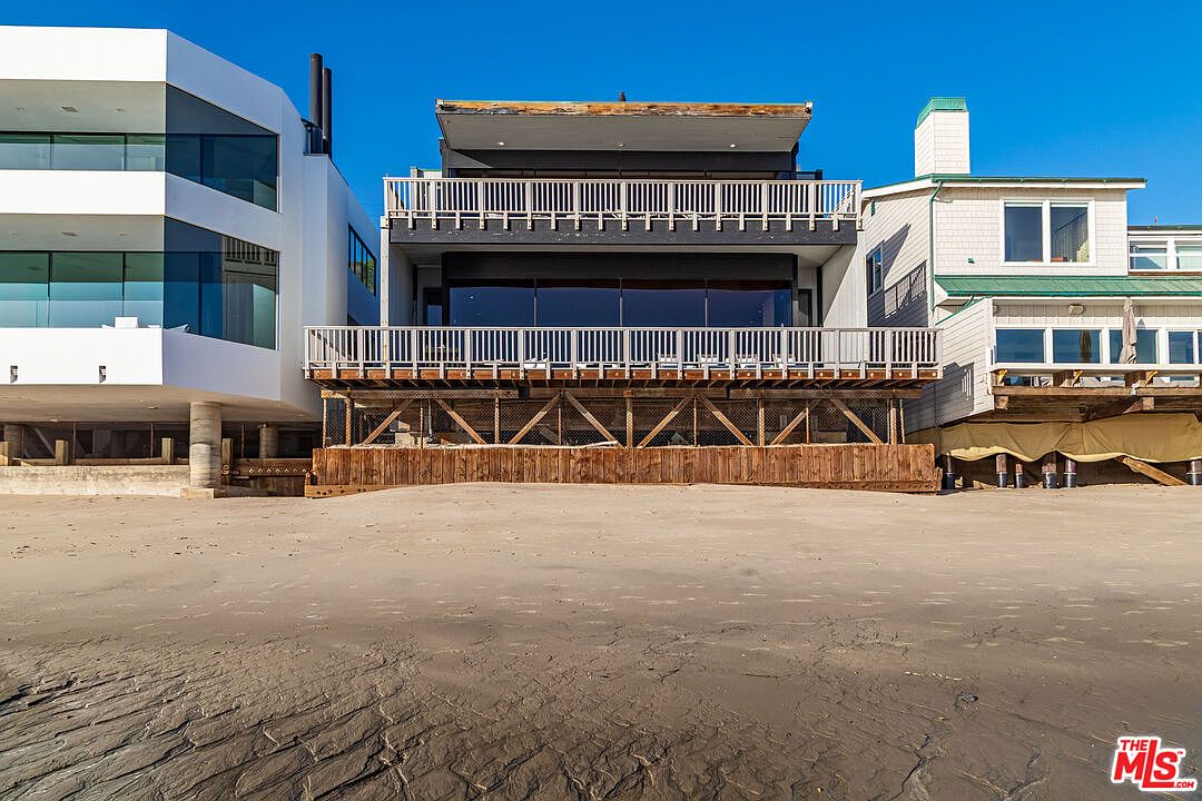 This is a rear view of a beachfront property showcasing multiple levels with balconies and large windows. The architecture includes a mix of modern and traditional elements, with wooden support structures visible beneath the building. The scene is set on a sandy beach under a clear blue sky, emphasizing the property's direct access to the ocean.