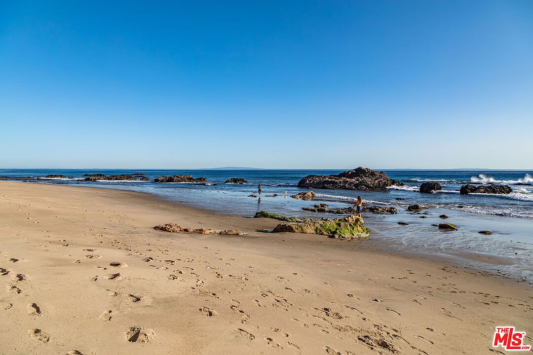 This image showcases a serene beach scene with a clear blue sky and calm ocean. Several rock formations are scattered along the shoreline, with gentle waves lapping at the sandy beach. The perspective is wide, capturing the vastness of the ocean and the tranquility of the beach, making it an inviting coastal property.