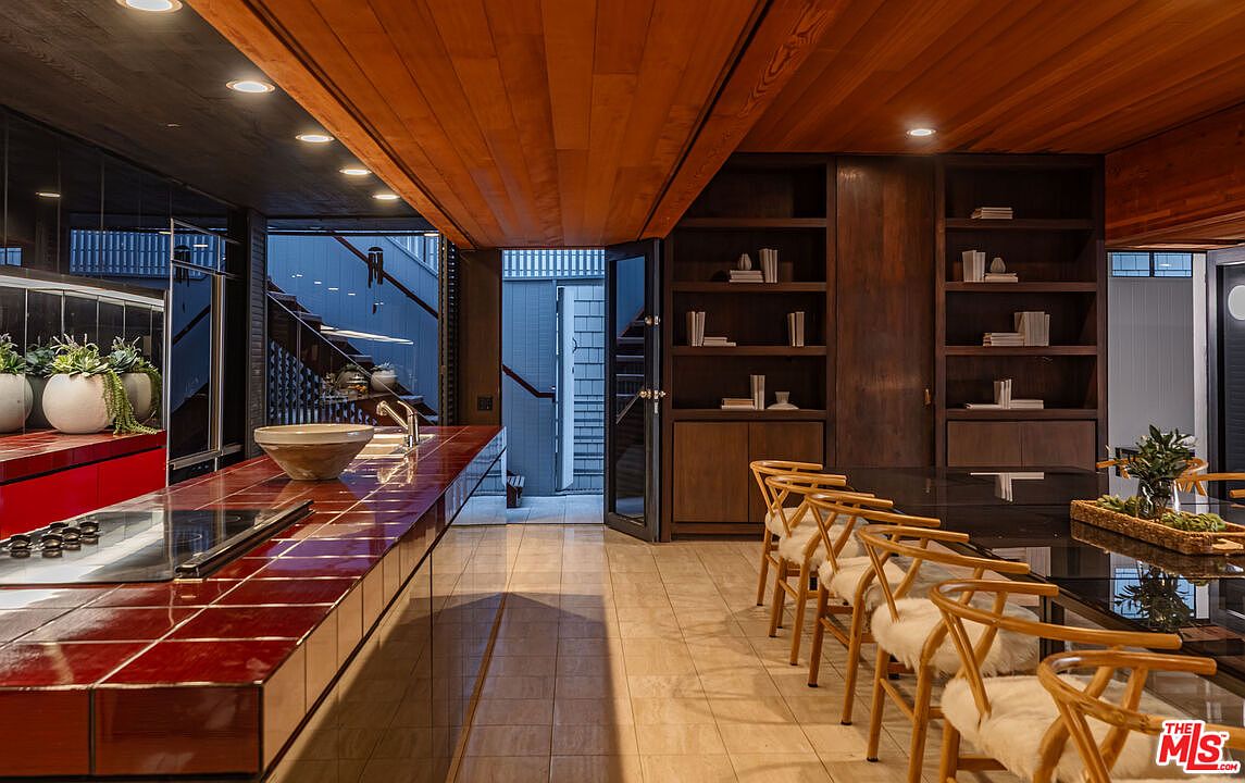 This interior shot showcases a modern kitchen with a unique design. The kitchen features a red tiled countertop, wooden ceiling, and dark wood cabinetry with open shelving. A dining area with wooden chairs and a black table is adjacent to the kitchen, creating an open and inviting space.