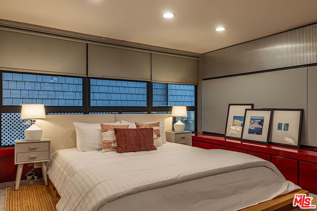 This is a primary bedroom featuring a large bed with white linens and decorative pillows. The room has a modern aesthetic with neutral tones, recessed lighting, and window shades. A red cabinet with framed artwork sits against one wall, and bedside tables with lamps flank the bed.