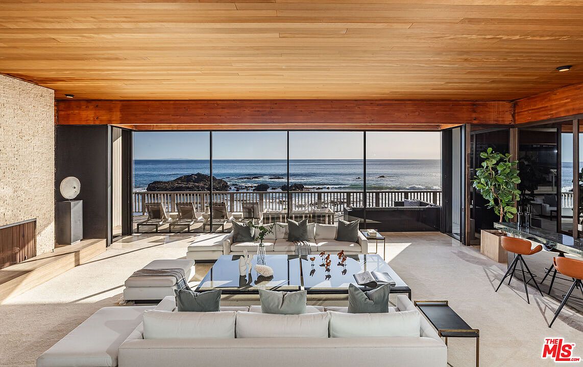 This is a luxurious living room with a stunning ocean view through expansive windows. The room features a large, white sectional sofa, a glass coffee table, and a warm wood ceiling, creating a bright and airy atmosphere. The design emphasizes a seamless connection between indoor and outdoor spaces, perfect for relaxation and entertainment.
