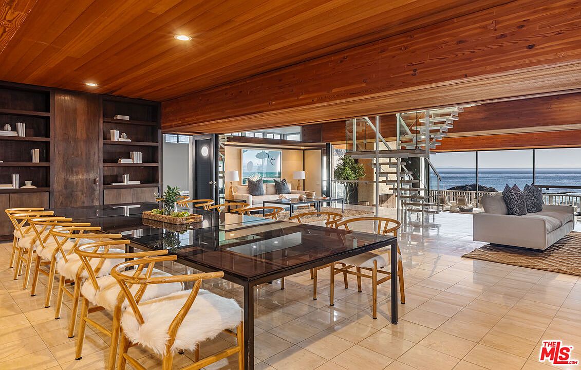 This is an interior shot of a dining room that opens to a living area with ocean views. The dining room features a large glass-topped table with wooden chairs and a built-in bookshelf. The living area has a modern staircase and a comfortable seating arrangement, with a large window showcasing the ocean.