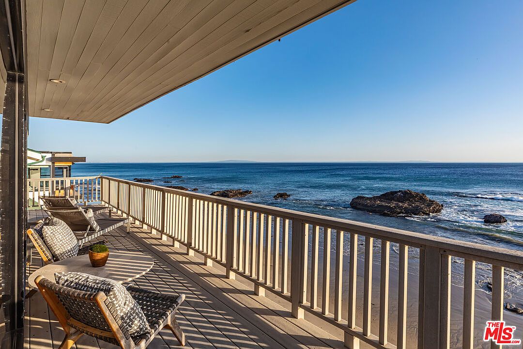 This image showcases a beautiful outdoor deck or balcony with stunning ocean views. The deck features comfortable seating arrangements, including chairs and a small table, perfect for relaxation. The wooden railing provides safety and enhances the coastal aesthetic, while the clear blue sky and ocean create a serene and inviting atmosphere.