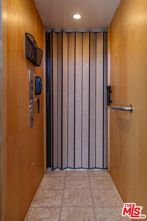 This interior shot showcases a residential elevator. The elevator features a folding door with vertical wood panels and black trim. The hallway has tiled flooring and wood paneled walls, with a control panel and a small wire basket mounted on one side. A handrail is visible on the right side of the elevator door.
