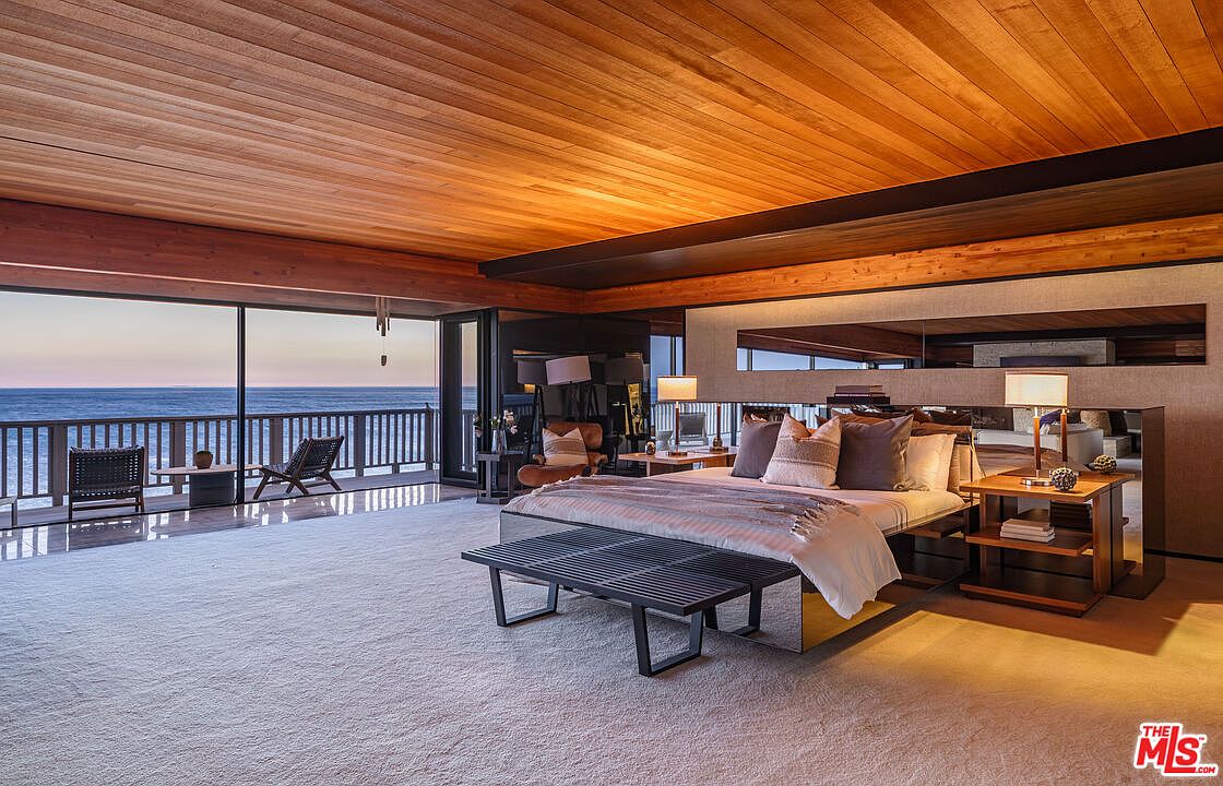 This is a luxurious primary bedroom with a stunning ocean view. The room features a wood-paneled ceiling, a large bed with multiple pillows, and a bench at the foot of the bed. Sliding glass doors lead to a balcony overlooking the ocean, creating a serene and inviting atmosphere.
