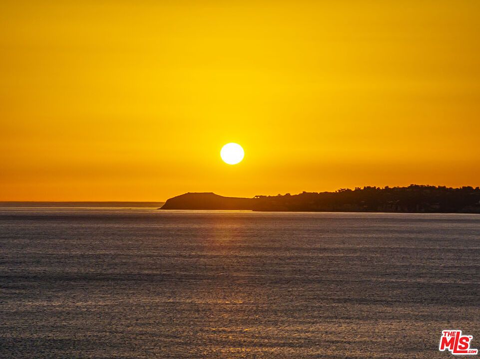 The image captures a serene sunset over the ocean, with the sun positioned prominently in the sky, casting a warm golden glow across the water and horizon. A silhouette of land is visible in the distance, adding depth to the scene. The overall impression is one of tranquility and natural beauty, highlighting the potential for stunning views from a property.