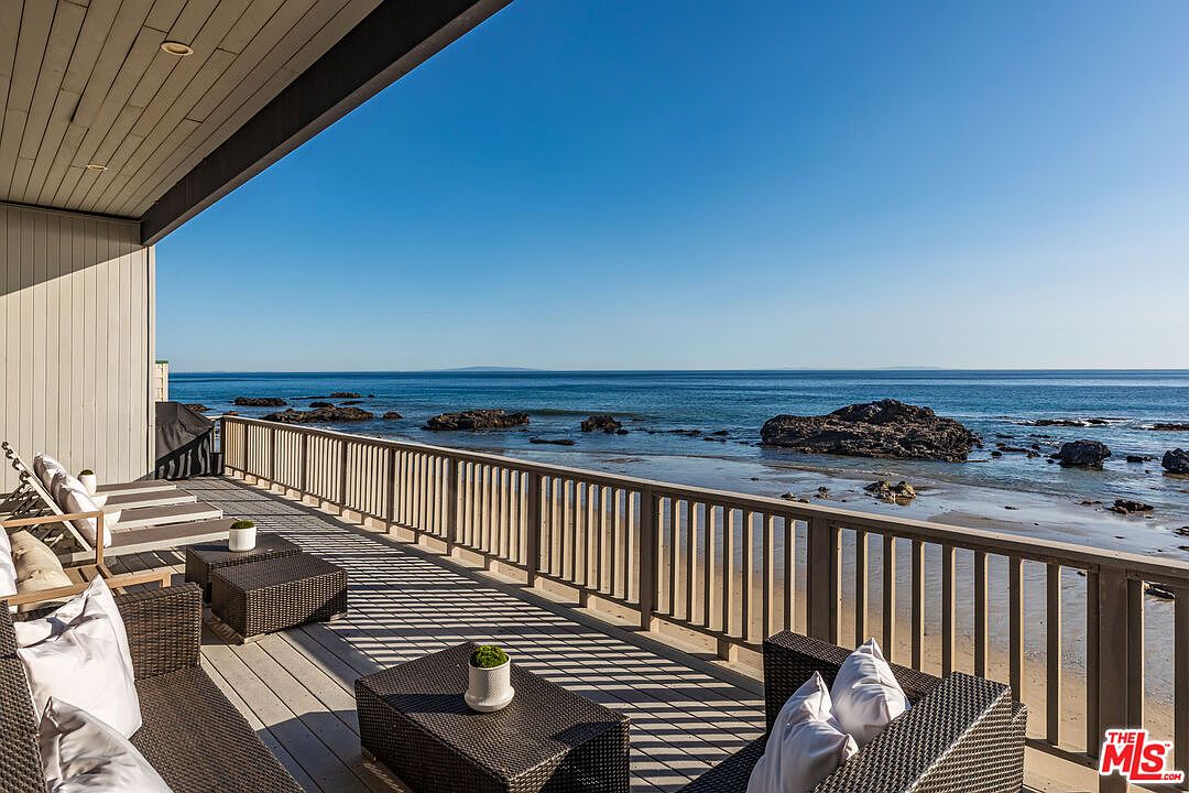 This image showcases a beautiful outdoor deck or balcony with stunning ocean views. The deck features comfortable wicker furniture with white cushions, perfect for relaxation. A wooden railing provides safety and enhances the coastal aesthetic, while the clear blue sky and ocean create a serene and inviting atmosphere.