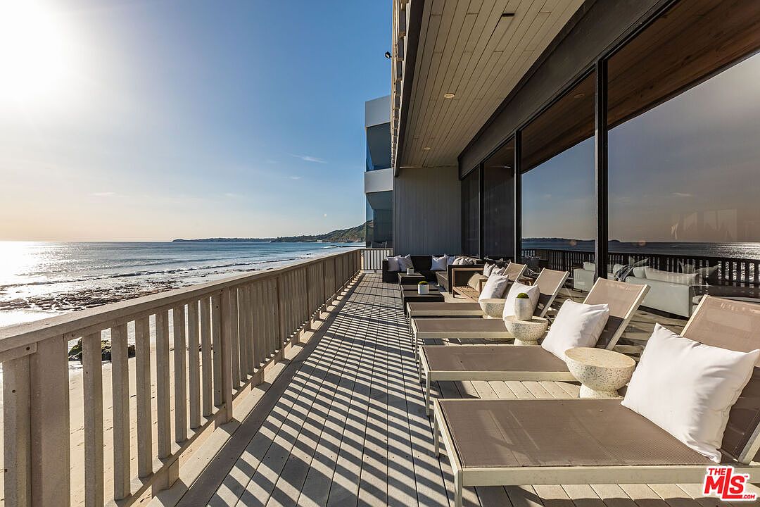 This image showcases a luxurious oceanfront patio or deck, featuring multiple lounge chairs with white cushions and small side tables. The deck is constructed with light-colored wood and has a sturdy railing, offering unobstructed views of the ocean and coastline. Large windows reflect the ocean view, suggesting a seamless indoor-outdoor living experience.