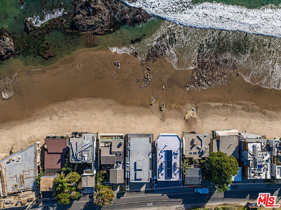 This aerial shot showcases a row of beachfront properties nestled along a sandy beach with waves gently crashing. The houses feature diverse architectural styles, with flat roofs and modern designs. A road runs parallel to the houses, providing access to the properties, while the ocean stretches out in the background.