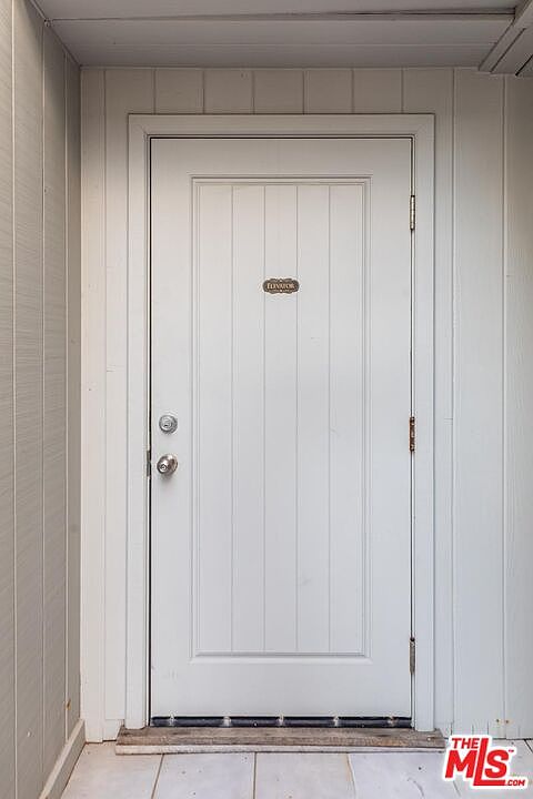 The image showcases a well-maintained entryway featuring a white, paneled front door. The door is framed by matching white trim and siding, creating a clean and inviting appearance. A small nameplate is visible above the doorknob, adding a touch of personalization. The perspective is a straight-on shot, emphasizing the door's central position.