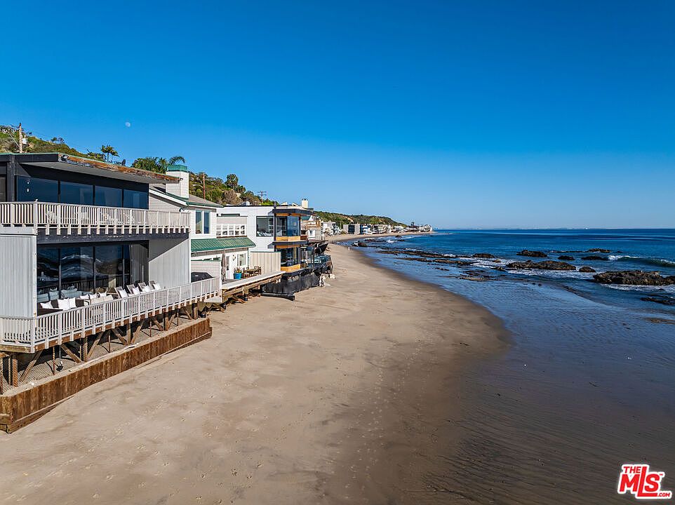This aerial view showcases a row of beachfront homes nestled along a sandy shore, with the vast expanse of the ocean stretching out to the horizon under a clear blue sky. The homes feature a mix of architectural styles, with decks and balconies overlooking the beach. The scene evokes a sense of luxury coastal living.