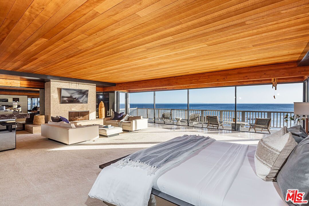 This is a luxurious primary bedroom with a stunning ocean view through floor-to-ceiling windows. The room features a spacious layout with a seating area including a fireplace and a large bed in the foreground. The wood-paneled ceiling adds warmth, while the neutral color palette creates a serene and inviting atmosphere.