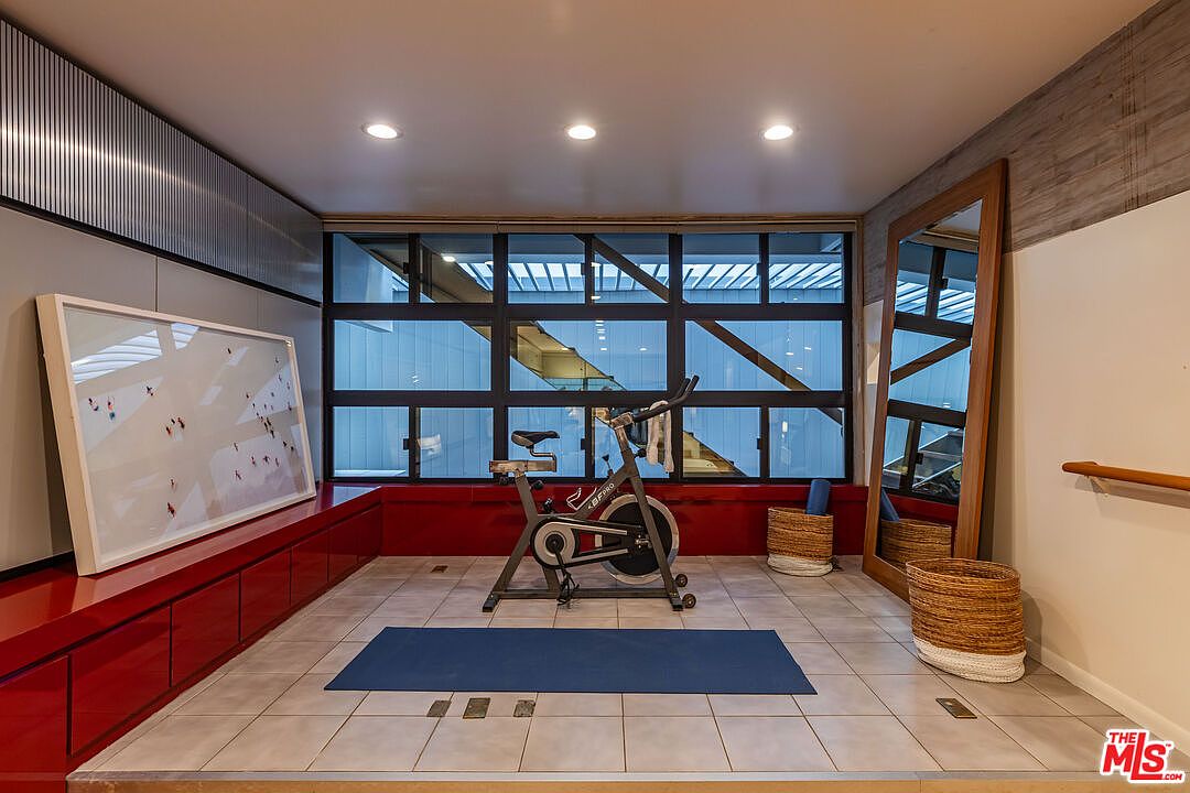 This is an interior shot of a home gym featuring a stationary bike on a blue yoga mat, positioned in front of a large window. The room includes built-in red cabinetry, a framed artwork, and a full-length mirror, creating a dedicated and stylish workout space. The tile flooring and recessed lighting contribute to a clean and functional environment.