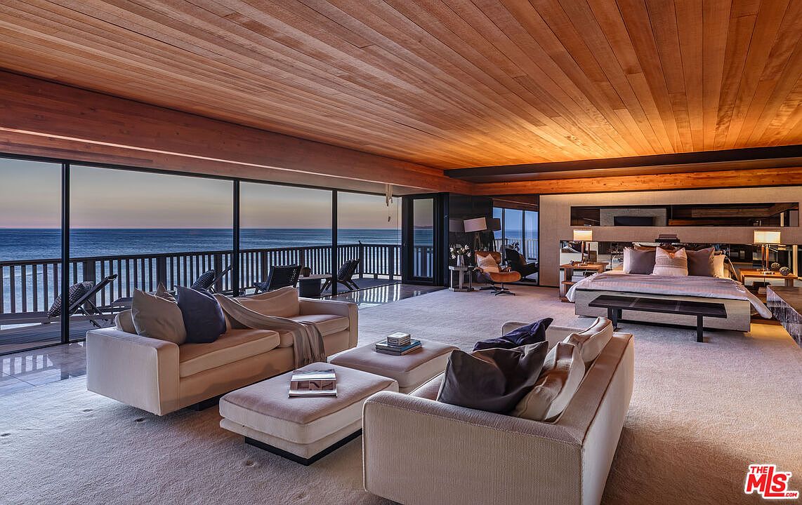 This is a luxurious primary bedroom with an open floor plan that seamlessly integrates a living area and sleeping space. The room features floor-to-ceiling windows offering ocean views, a wood-paneled ceiling, and contemporary furnishings including two sofas, ottomans, and a king-size bed with a bench. The overall impression is one of spaciousness, elegance, and tranquility.