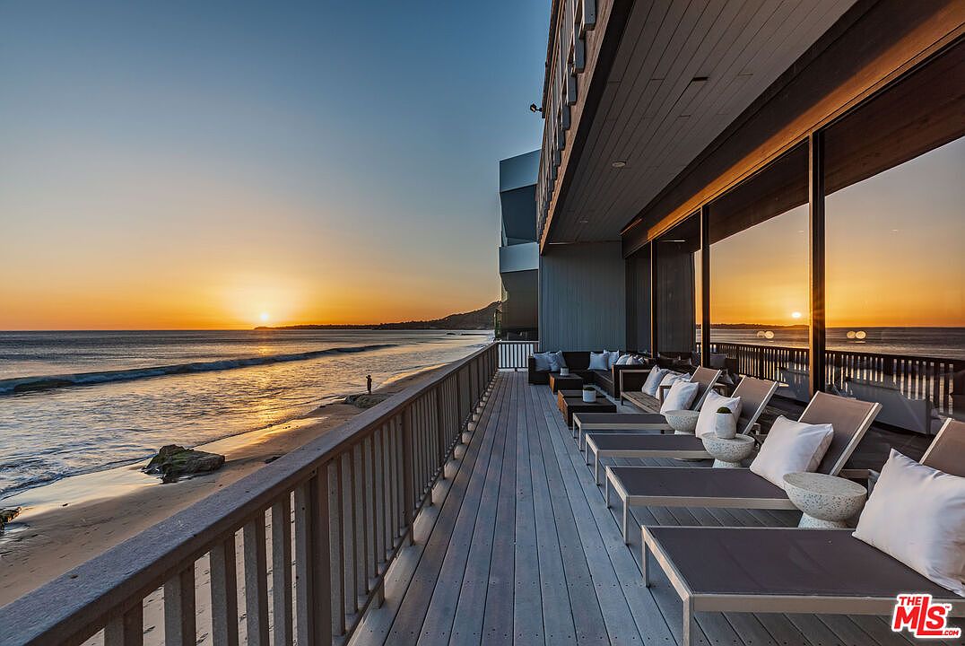 This image showcases a luxurious beachfront patio or deck at sunset. The space features multiple lounge chairs with white cushions and small side tables, offering a relaxing area with ocean views. The building's architecture includes large windows that reflect the sunset, and a wooden railing provides safety along the beachside edge.