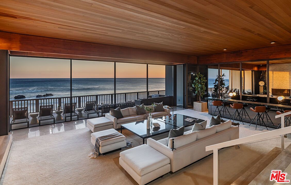 This is a luxurious living room with a stunning ocean view through large windows. The room features a spacious seating area with modern sofas, a glass coffee table, and a neutral-toned rug. The wood-paneled ceiling adds warmth, while the adjacent bar area with stools provides an entertainment space, creating an inviting and sophisticated atmosphere.