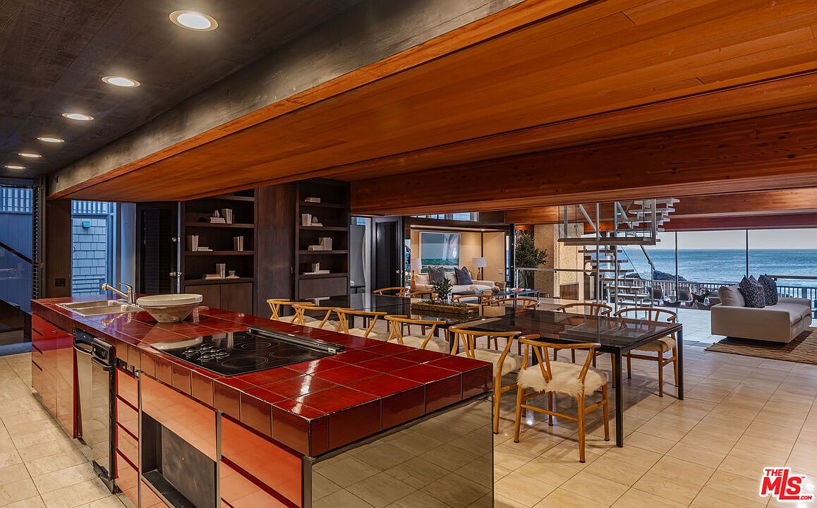 This interior shot showcases a modern kitchen and dining area with an open floor plan, featuring a red-tiled kitchen island with a sink and cooktop, wooden cabinetry, and a dining table with unique chairs. Large windows offer stunning ocean views, and a spiral staircase adds architectural interest. The space is well-lit and designed for both functionality and aesthetic appeal.