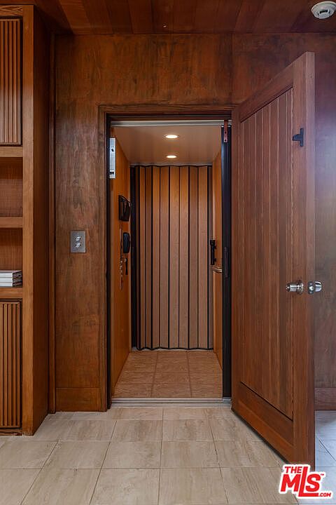 The image showcases an interior hallway featuring an elevator with wooden paneling. The elevator door is partially open, revealing a vertically paneled interior. The hallway is finished with light-colored tile flooring and matching wooden wall panels, creating a warm and classic aesthetic.