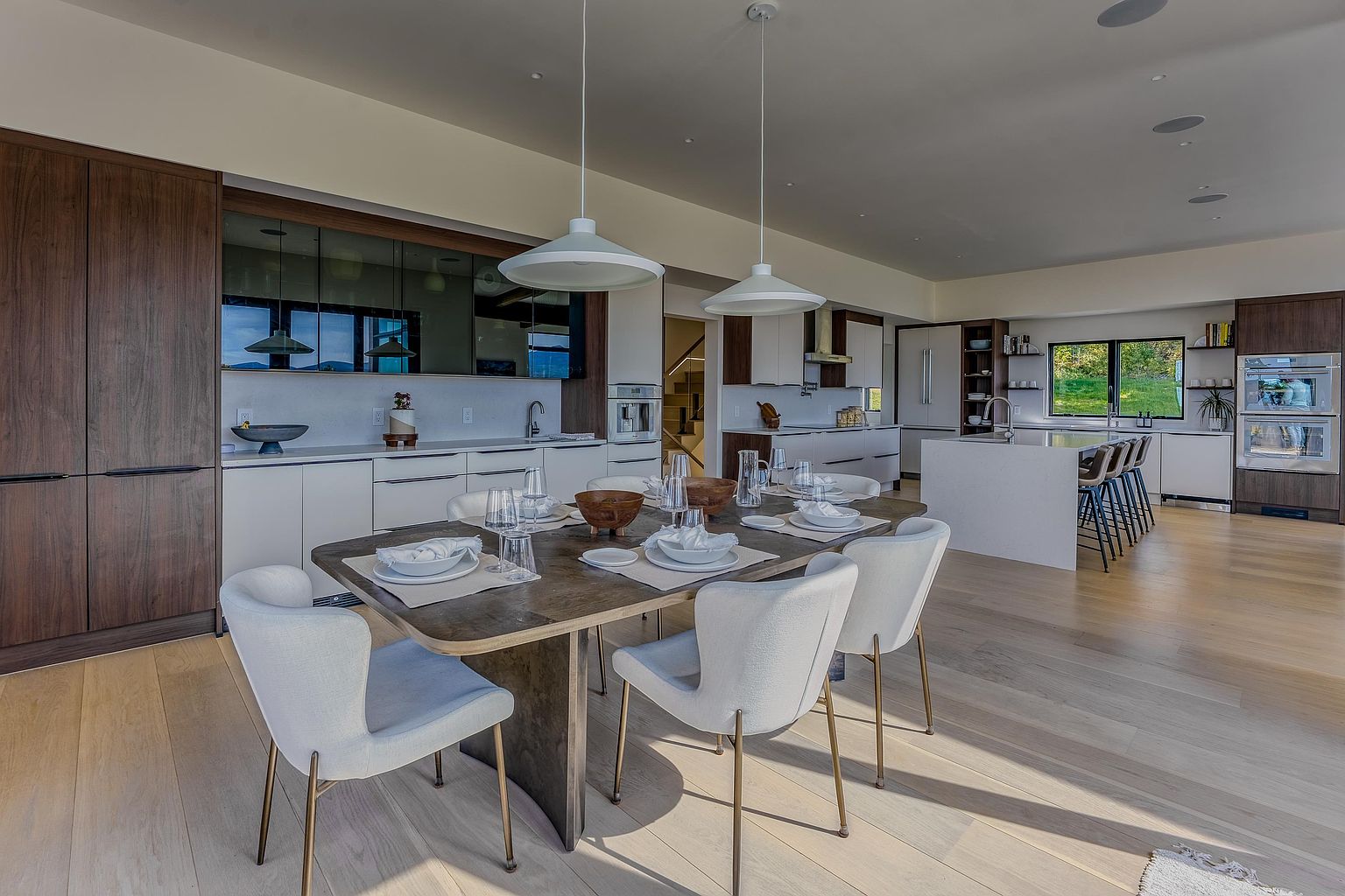 This expansive, open-concept kitchen and dining area features a sophisticated blend of warm wood cabinetry and sleek white finishes. A large, central dining table with upholstered chairs anchors the space, while the kitchen boasts a spacious island with bar seating and high-end stainless steel appliances. The room is bathed in natural light, creating an inviting and modern atmosphere perfect for both daily living and entertaining.
