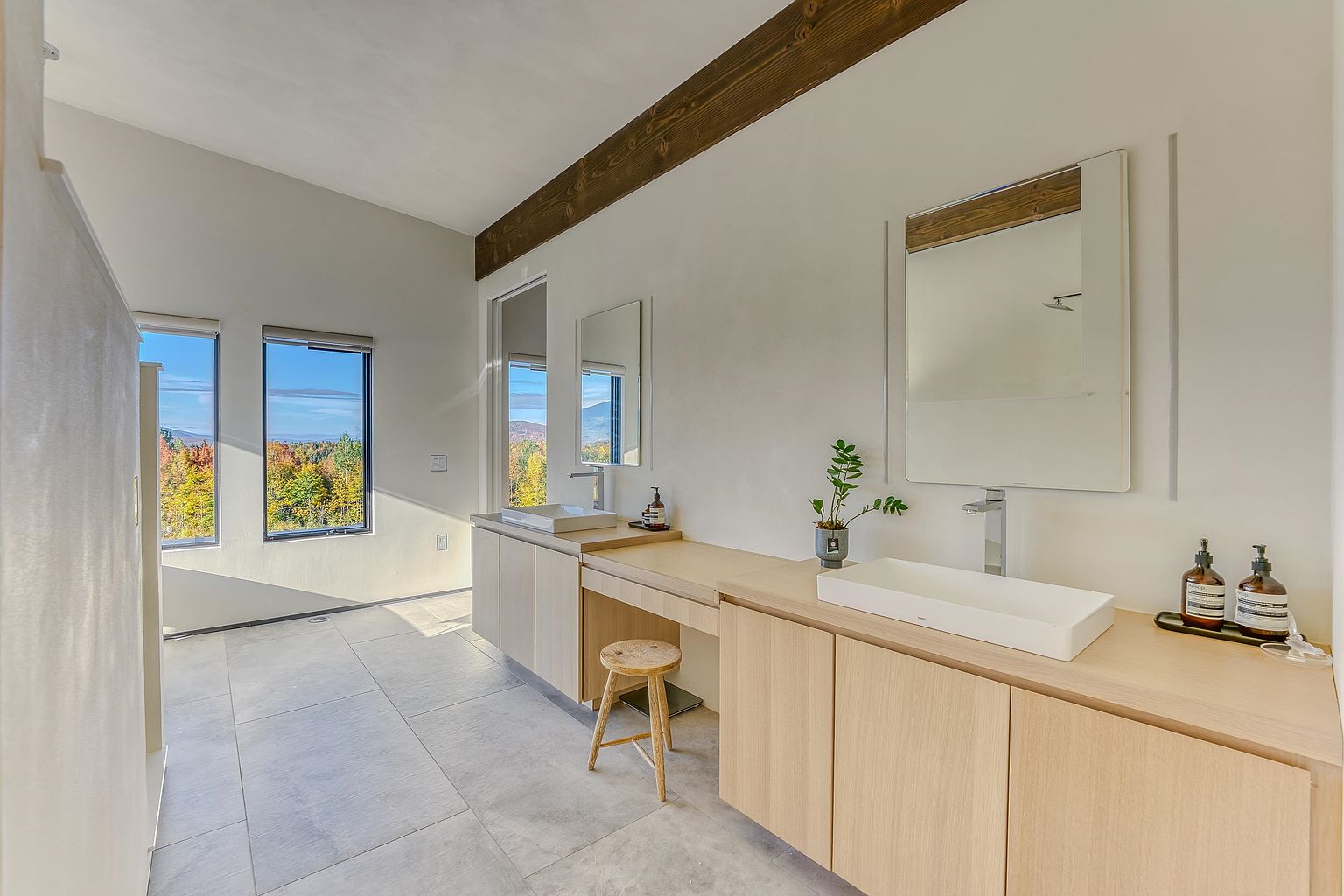 This modern primary bathroom features a sleek, minimalist design with light wood cabinetry, dual vessel sinks, and a dedicated vanity area with a wooden stool. Large-format grey floor tiles and a high ceiling with a rustic wooden beam create a sophisticated, airy atmosphere, while expansive windows offer a stunning view of the surrounding autumn forest. The perspective is a wide-angle shot that captures the clean lines and serene, spa-like aesthetic of the space.