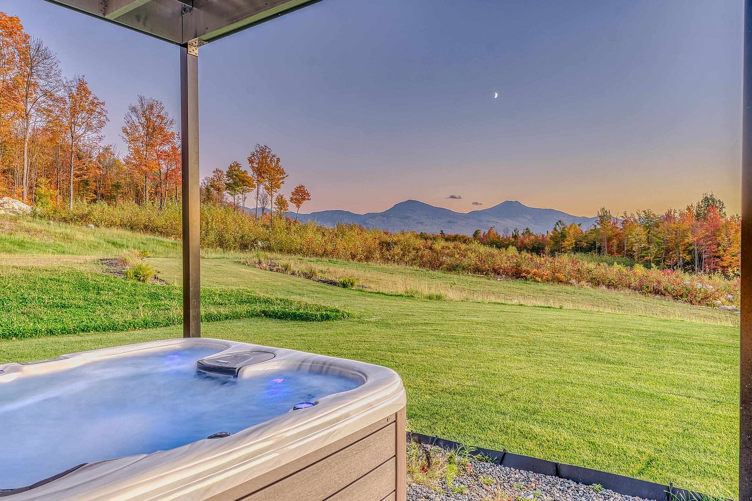 This image captures a serene outdoor scene from a covered patio, featuring a modern hot tub with glowing blue water as the primary focal point. Beyond the patio, a lush green lawn stretches toward a vibrant autumn forest and majestic mountain range under a clear twilight sky with a visible crescent moon. The perspective offers a relaxing, cinematic view that emphasizes the property's tranquil atmosphere and scenic natural surroundings.
