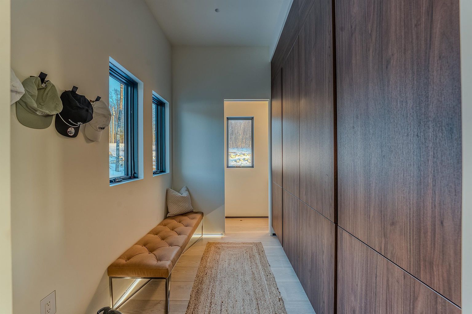 This modern, minimalist hallway features a long, tufted leather bench set against a wall with two windows, creating a bright and inviting transition space. A full-length wall of dark wood cabinetry provides extensive storage, while a textured runner rug adds warmth to the light-toned flooring. The perspective looks down the corridor toward a doorway, highlighting the clean lines and sophisticated, contemporary design.