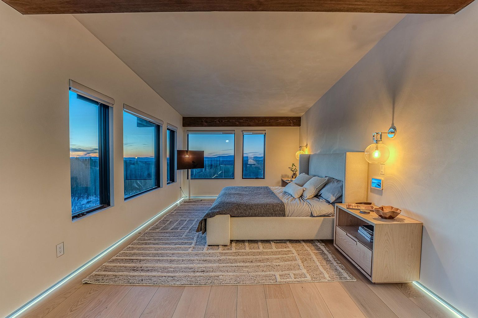 This modern primary bedroom features a minimalist aesthetic with a neutral color palette, highlighted by a plush upholstered bed and a textured area rug. Large windows frame a stunning sunset view, while warm wall-mounted globe lighting and under-bed LED accent lighting create a cozy, sophisticated atmosphere. The room is anchored by a sleek wooden nightstand, emphasizing a clean, contemporary design perfect for a luxury retreat.