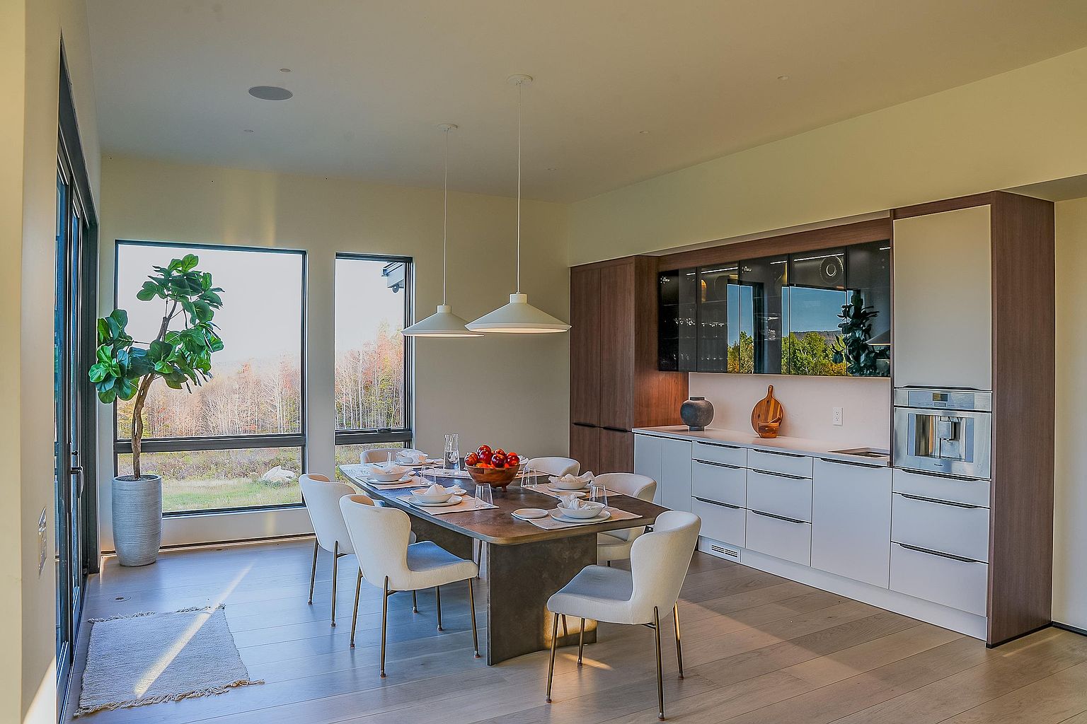This elegant dining area seamlessly integrates with a modern kitchen, featuring a sleek, dark-toned dining table surrounded by contemporary upholstered chairs. Large floor-to-ceiling windows flood the space with natural light and offer expansive views of the surrounding landscape, while minimalist pendant lights hang above the table. The room's sophisticated design is complemented by high-end cabinetry and a built-in oven, creating a cohesive and luxurious open-concept living space.