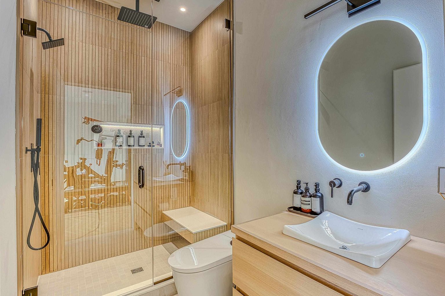 This modern, spa-inspired bathroom features a sleek glass-enclosed walk-in shower with vertical wood-slat tiling and a built-in bench. A contemporary vessel sink sits atop a light wood vanity, complemented by a backlit oval mirror and minimalist wall-mounted fixtures. The overall aesthetic is warm, sophisticated, and highly curated, emphasizing clean lines and high-end materials.