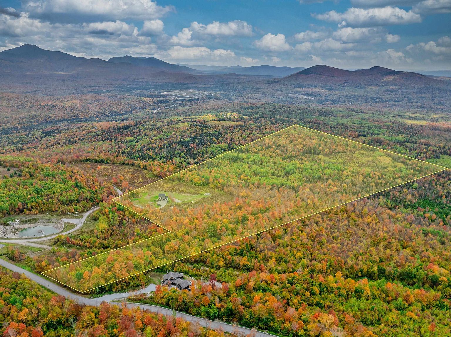 This high-altitude aerial shot captures a vast, wooded property in a mountainous region during peak autumn foliage. A yellow outline highlights the expansive land parcel, which includes a small clearing with a structure and a larger, more developed home nestled near the road at the bottom of the frame. The dramatic backdrop of rolling mountains under a cloudy sky emphasizes the property's secluded and scenic location.