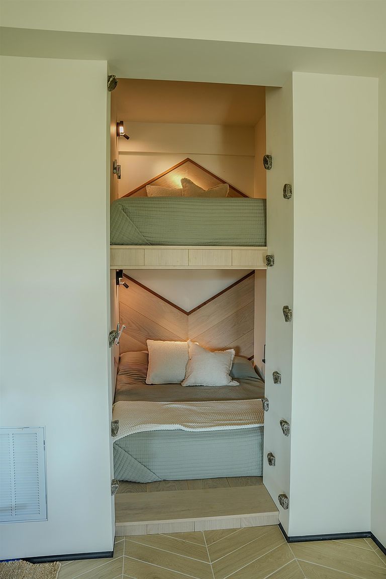 This image showcases a space-efficient, built-in bunk bed alcove featuring a modern, minimalist design with light wood accents and chevron-patterned headboards. The cozy nook includes integrated lighting for both the upper and lower bunks, creating a warm and inviting atmosphere. The perspective is a straight-on, eye-level shot that highlights the clever use of vertical space in a compact bedroom setting.