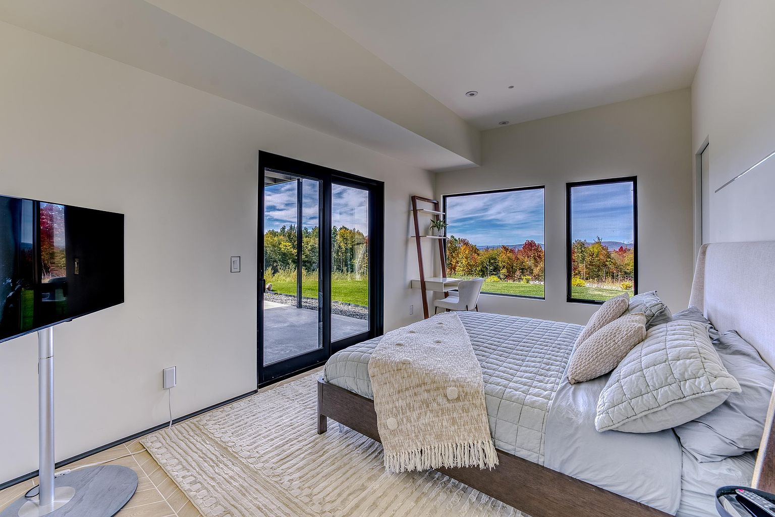 This modern primary bedroom features a minimalist aesthetic with a neutral color palette, centered around a plush bed dressed in soft linens and a textured throw. Large floor-to-ceiling windows and a sliding glass door flood the space with natural light, offering a stunning, unobstructed view of the surrounding autumn landscape. A sleek desk area tucked by the window and a wall-mounted television complete this serene and functional retreat.