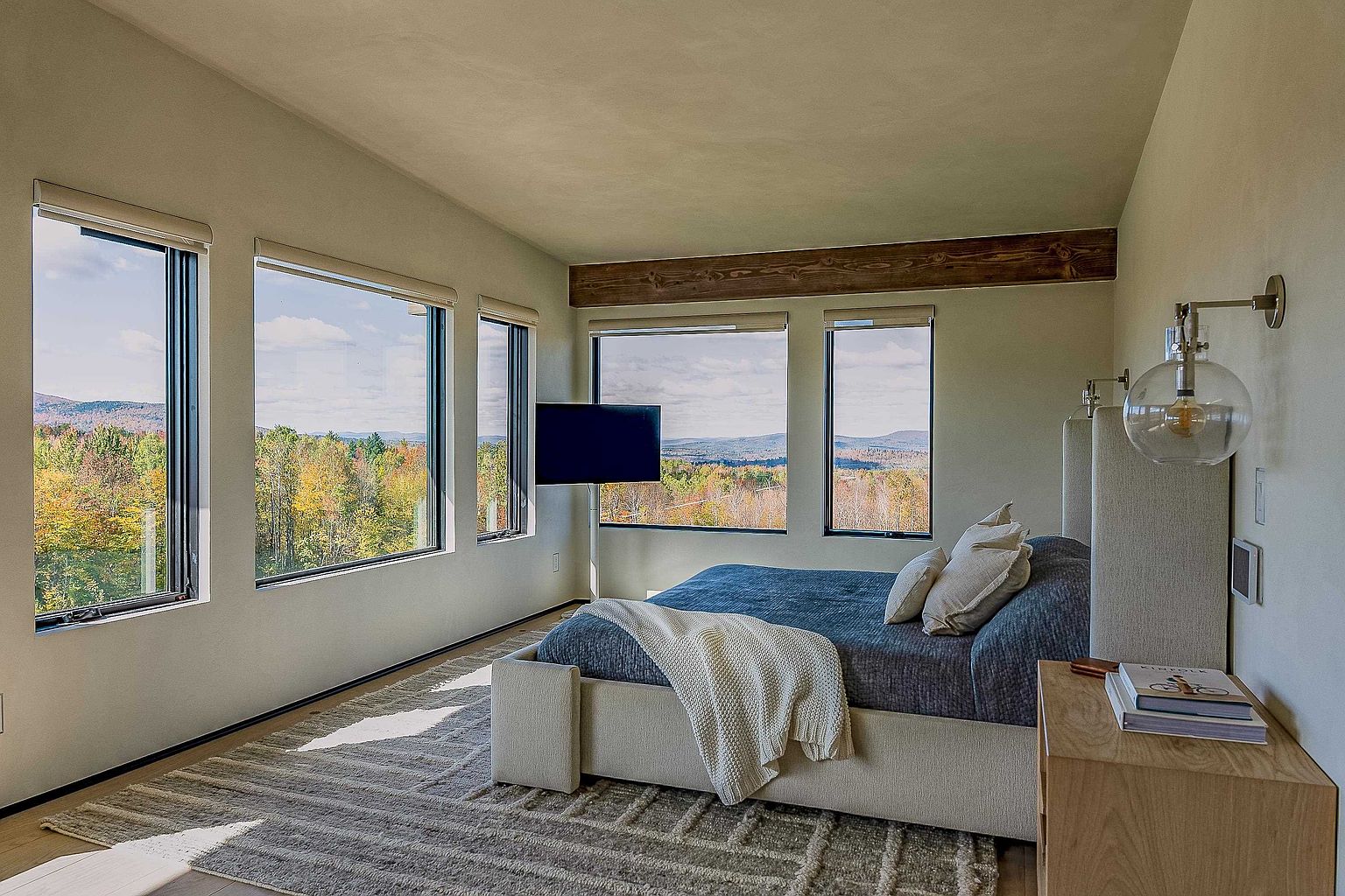 This serene primary bedroom features a minimalist, modern aesthetic with a plush bed centered against a backdrop of expansive windows that frame a breathtaking panoramic view of a forested mountain landscape. A rustic wooden ceiling beam adds warmth to the neutral color palette, while a sleek wall-mounted television and elegant glass globe sconces enhance the sophisticated, tranquil atmosphere. The room is anchored by a textured area rug, creating a cozy and inviting retreat that seamlessly blends indoor comfort with the natural beauty outside.
