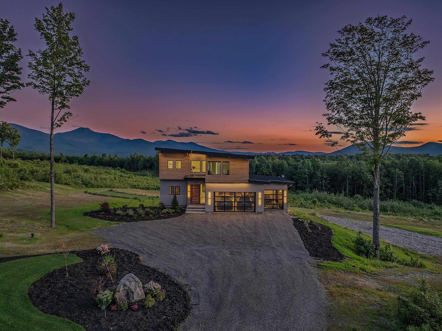 This stunning aerial view captures a modern, two-story home nestled against a backdrop of rolling mountains during a vibrant sunset. The property features a contemporary design with wood siding, large glass garage doors, and a spacious gravel driveway leading up to the entrance. The surrounding landscape includes manicured garden beds, mature trees, and expansive open space, creating a serene and picturesque setting.