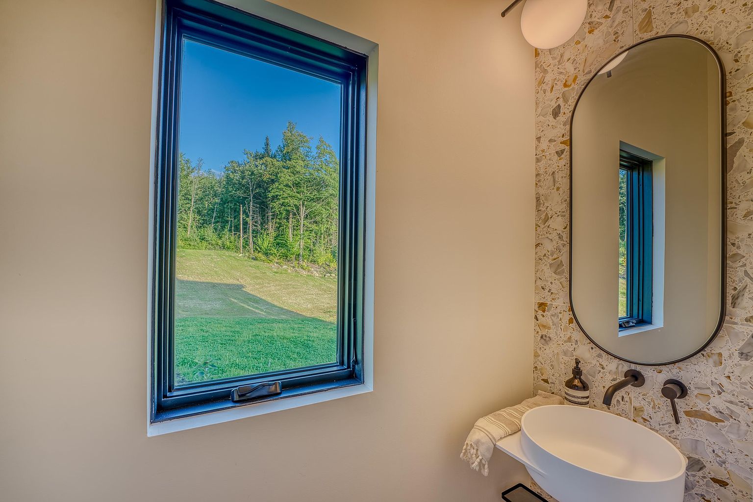 This modern powder room features a minimalist vessel sink mounted on a sleek shelf against a striking terrazzo-tiled wall. An oval mirror reflects the view from the adjacent window, which looks out onto a lush green landscape, creating a bright and airy atmosphere. The combination of natural textures and clean lines gives the space a contemporary, high-end feel.
