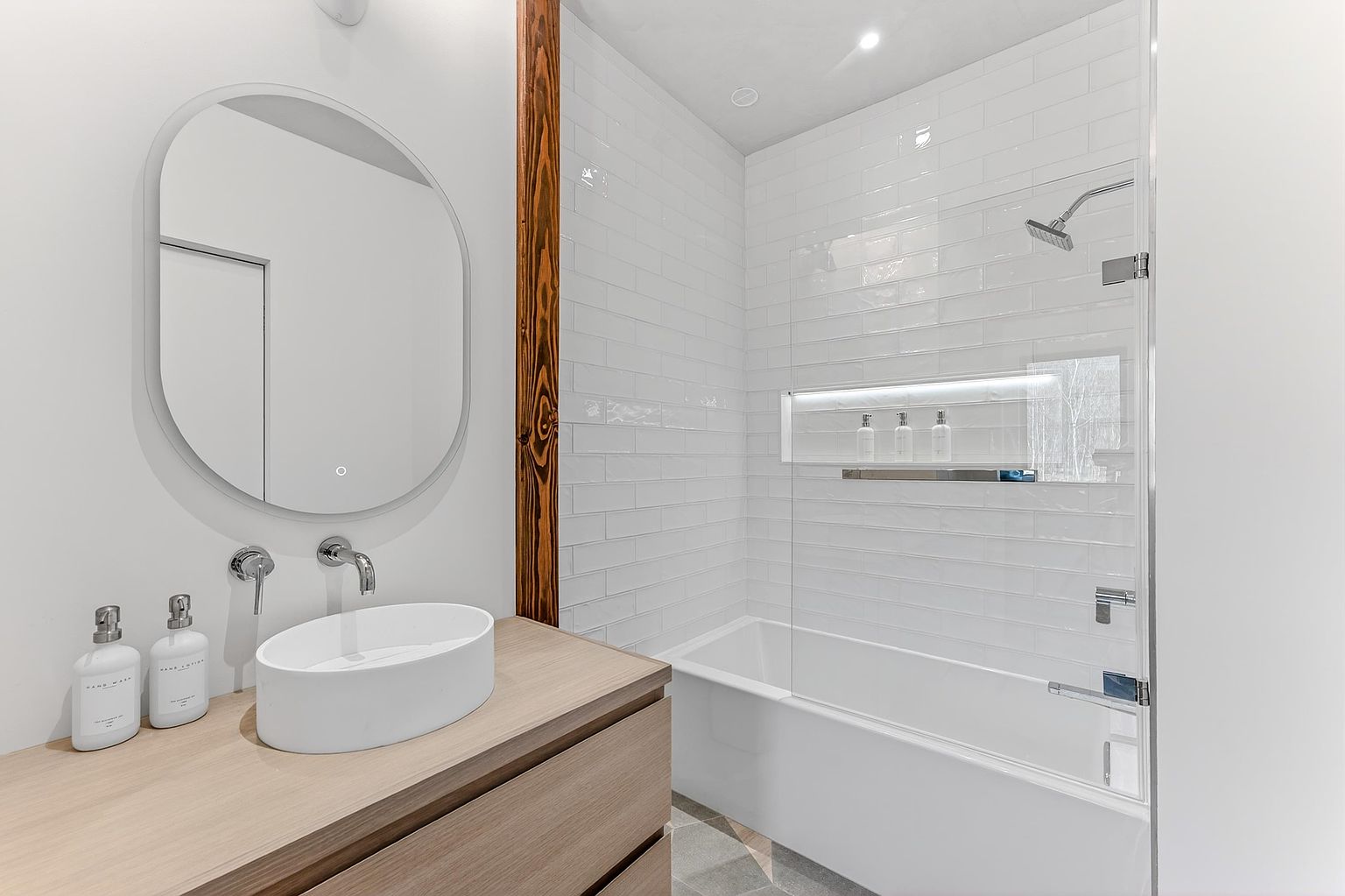 This modern, minimalist bathroom features a sleek vessel sink atop a light wood vanity, paired with a contemporary oval mirror. The space includes a clean, white-tiled shower-tub combination with a built-in, illuminated recessed shelf for storage. The overall aesthetic is bright, airy, and sophisticated, emphasizing high-end finishes and clean lines.